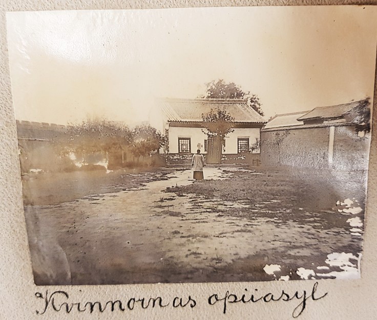 A sepia-toned photograph of a woman standing in front of a house with a garden, surrounded by greenery and other buildings.