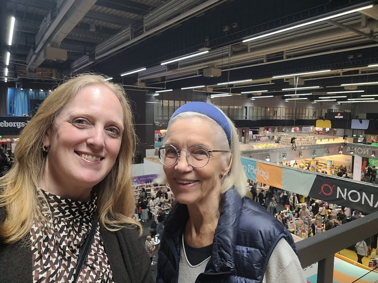 Two women smiling for a selfie indoors, overlooking a busy market or event space filled with stalls and visitors.