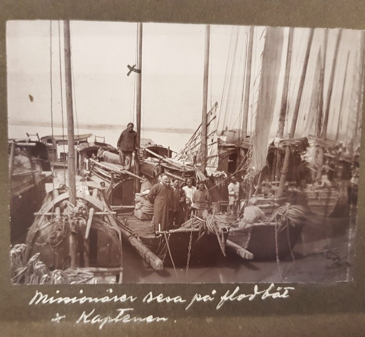 Historical photograph of several people on wooden boats at a dock, with tall sails and cargo visible, set against a river backdrop.