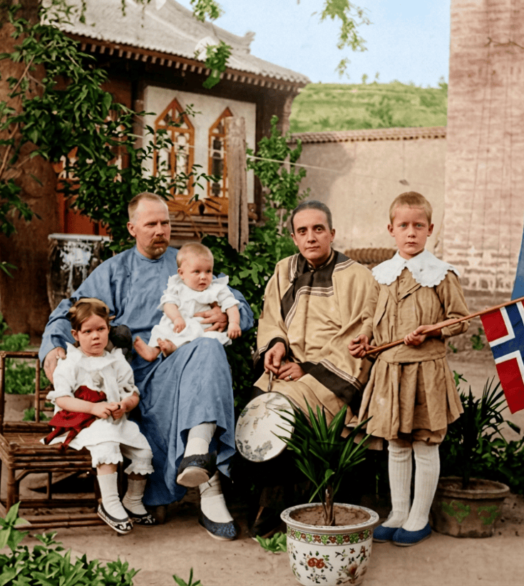 A family portrait featuring five individuals, including three children and two adults, seated amidst lush greenery and a decorative pot. The adults are dressed in traditional attire, while the children wear vintage clothing, one holding a flag.