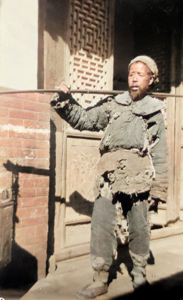 A man in tattered clothing holds a long pole, standing in front of an intricately carved wooden door.