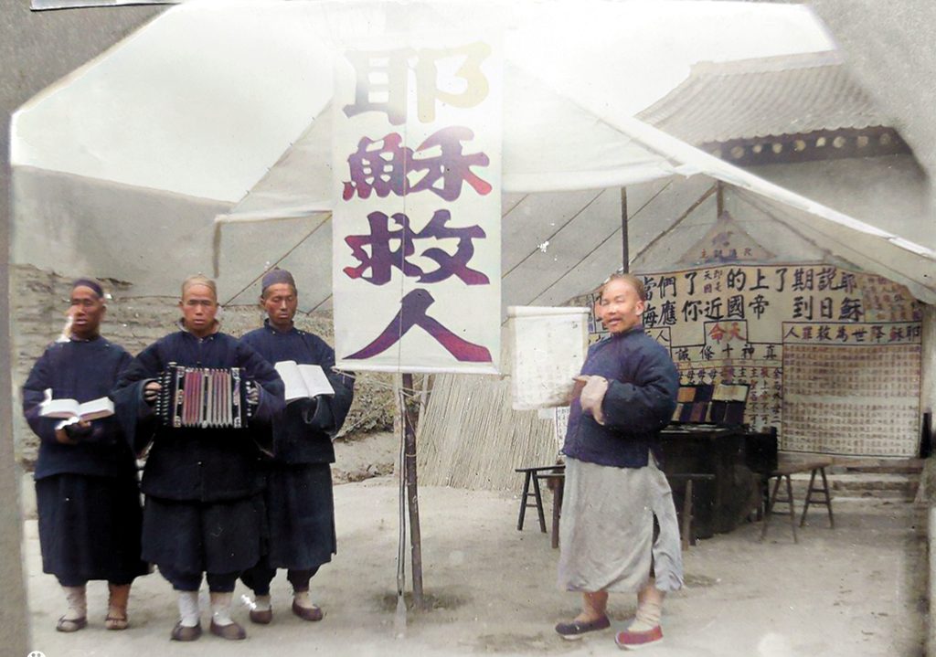 A historical scene featuring four men in traditional attire standing outside a tent. One man is holding a book, another is playing an accordion, and a third displays a sign. The tent has banners with calligraphy and other written content in the background.