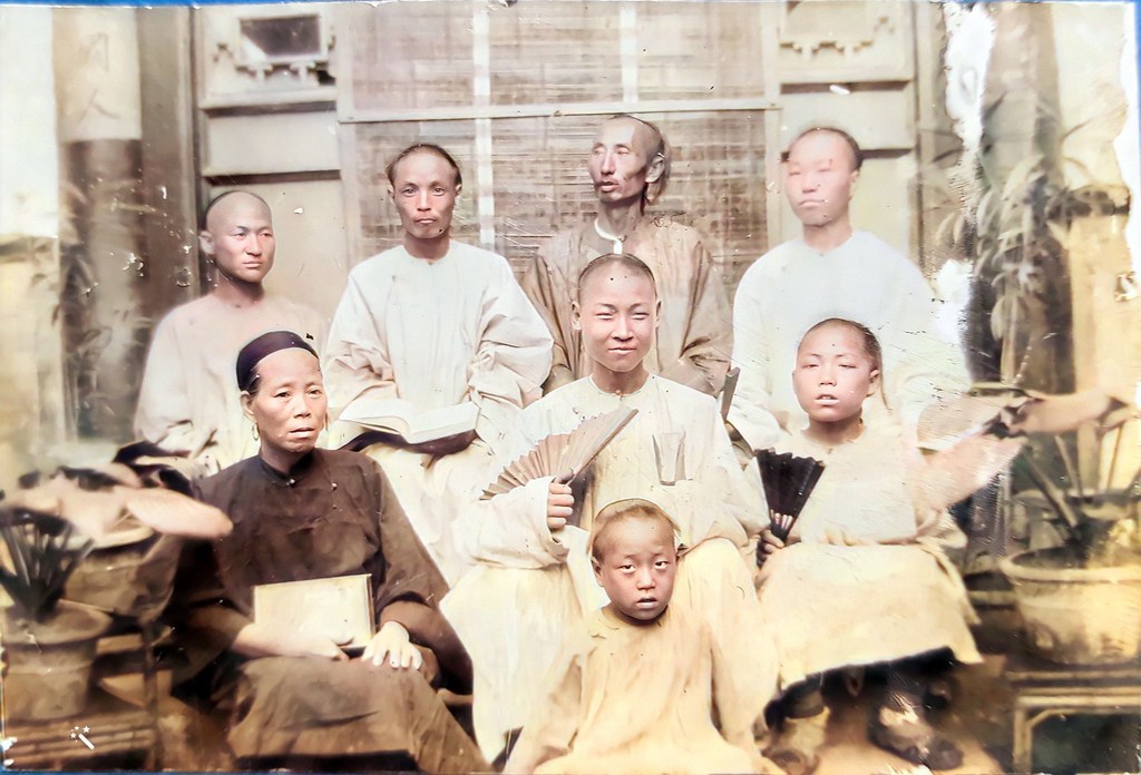 A historical portrait featuring a group of individuals sitting and standing together, with seven adults and two children, all with shaved heads. The adults are wearing traditional clothing, and some are holding books and fans. The setting is indoors, with visible plants in the background.