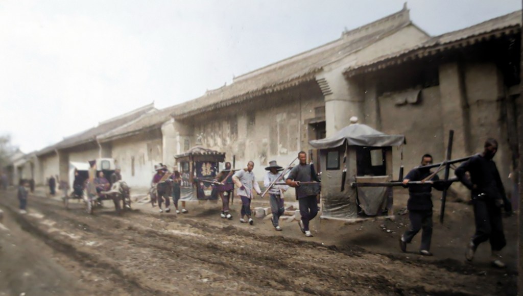 A group of men carrying traditional sedan chairs along a muddy street lined with rustic buildings, showcasing historical transportation methods.